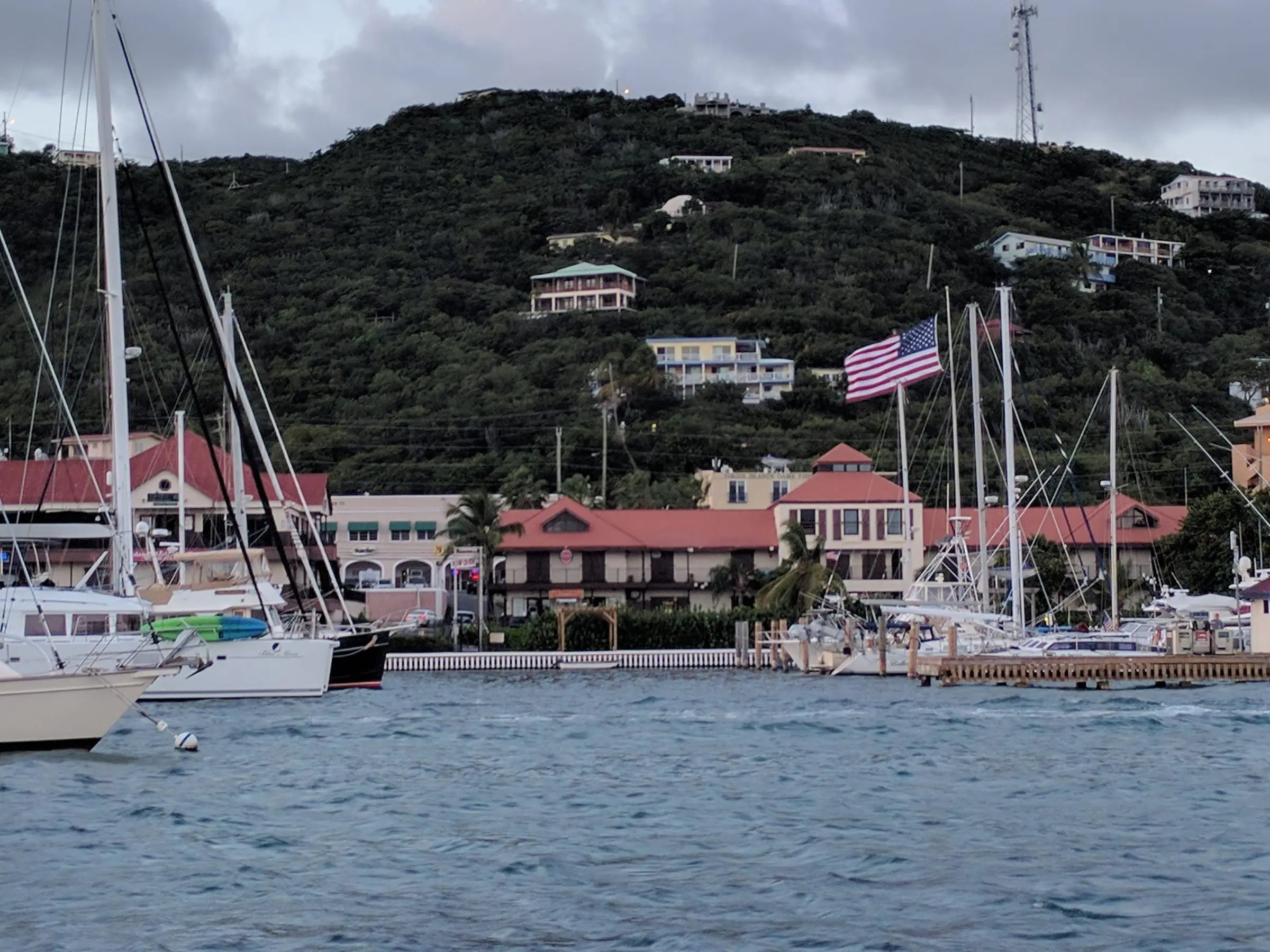 Red-roofed waterfront buildings of American Yacht Harbor behind its docks and moored sailboats on Red Hook Bay, with a large U.S. flag and hillside homes above