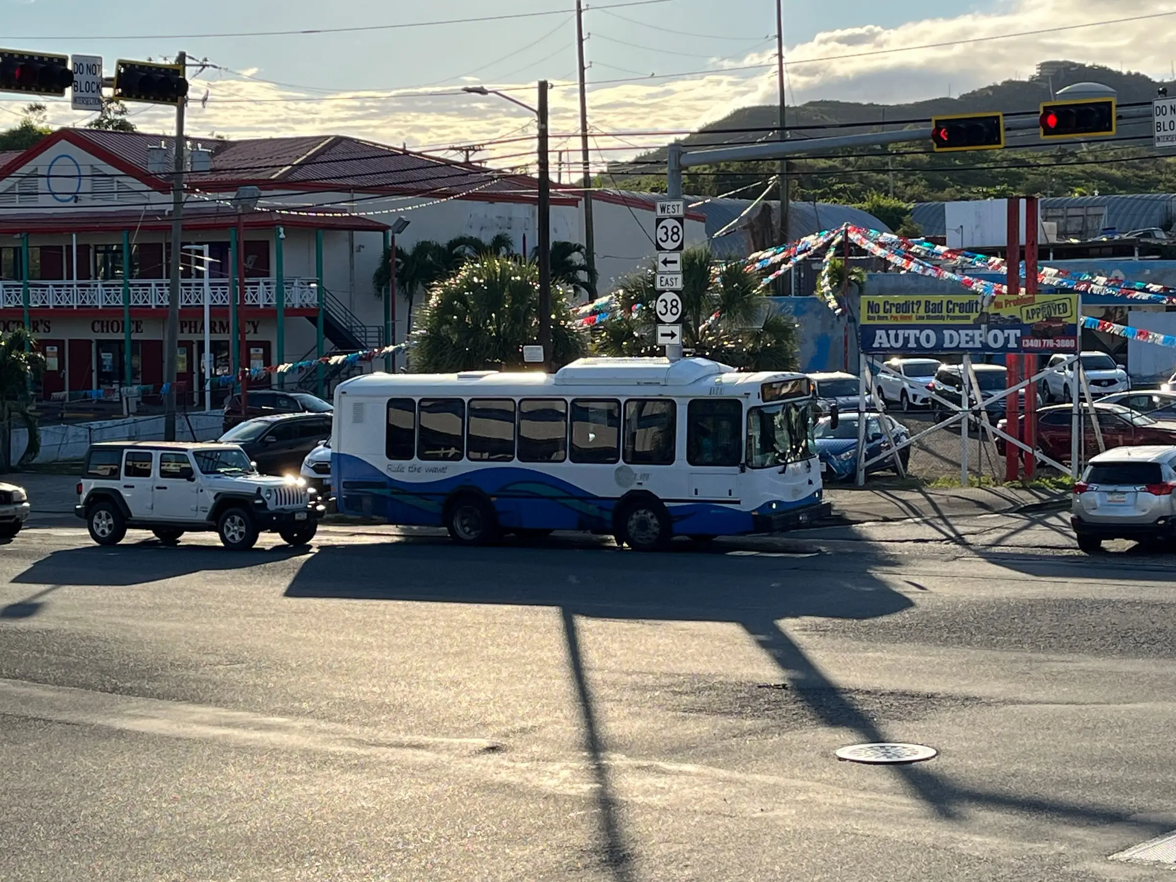 A white and blue VITRAN public bus crossing a Route 38 intersection in Anna's Retreat, with a pharmacy building and green hills behind