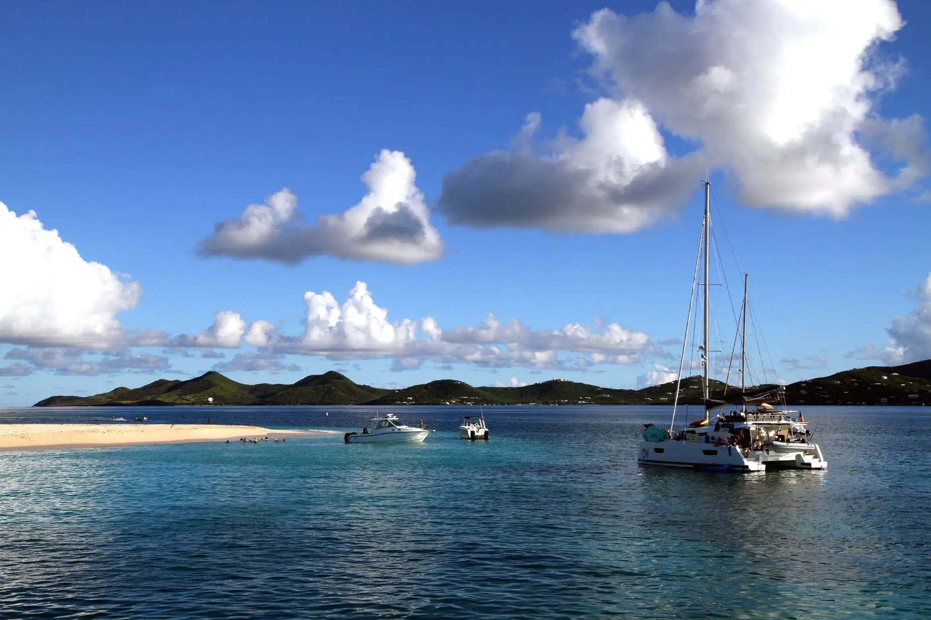 White sand spit at the west end of Buck Island with swimmers in the shallows and a catamaran and motorboats anchored offshore, the green hills of St. Croix's East End across the water
