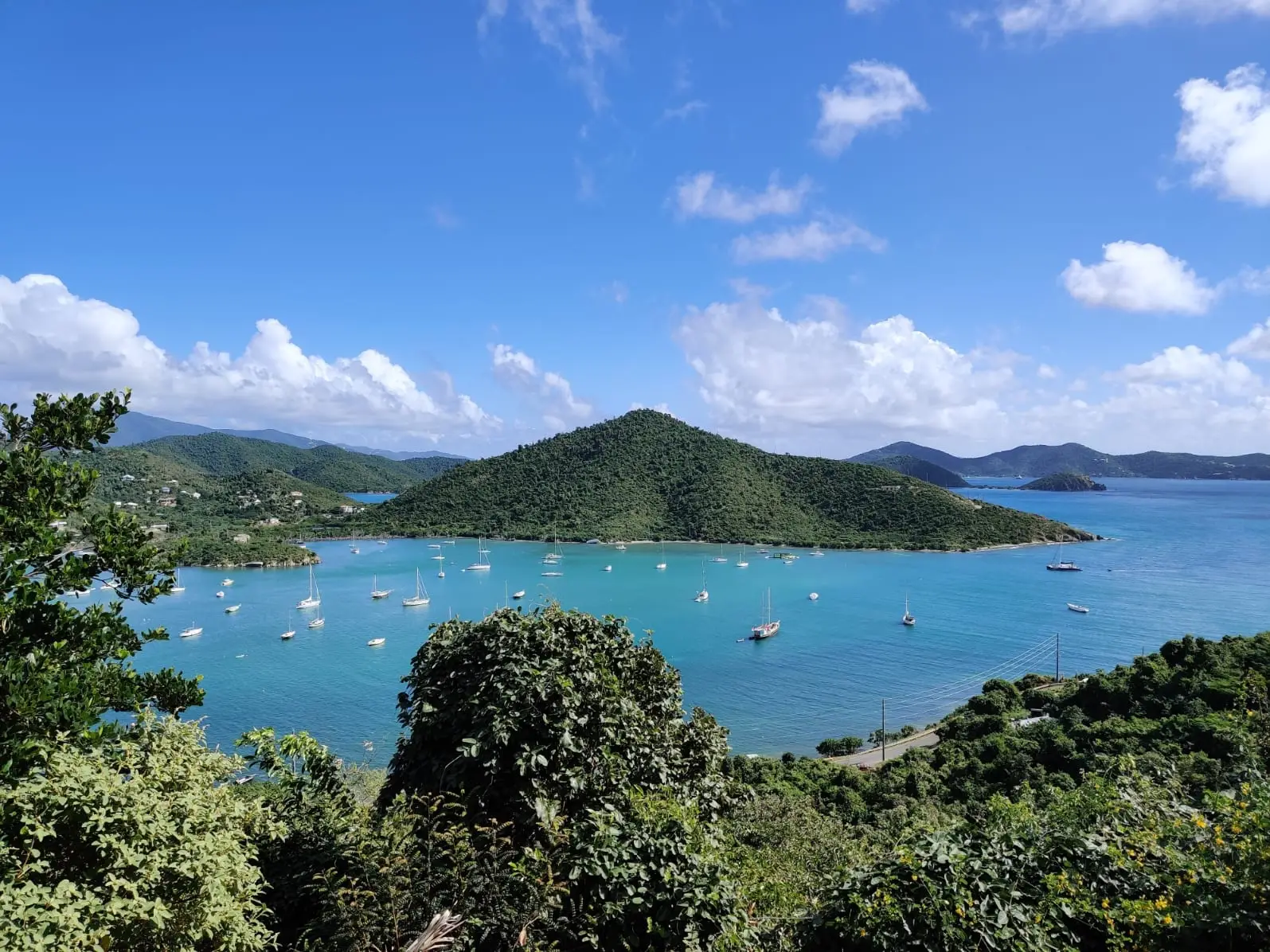 Sailboats moored across the turquoise water of Coral Bay below the green, cone-shaped hill of Fortsberg, with more islands on the horizon