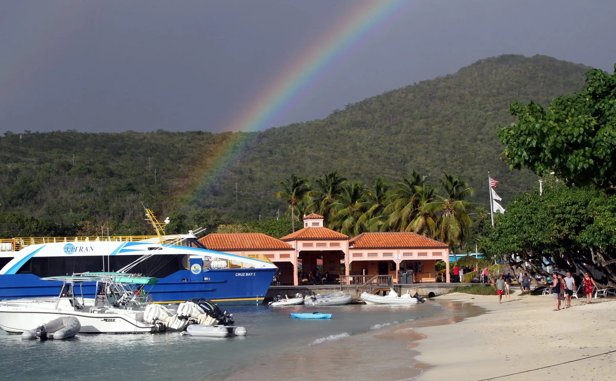 A VITRAN passenger ferry docked beside the orange-roofed ferry terminal and beach in Cruz Bay, with a rainbow over the green hillside