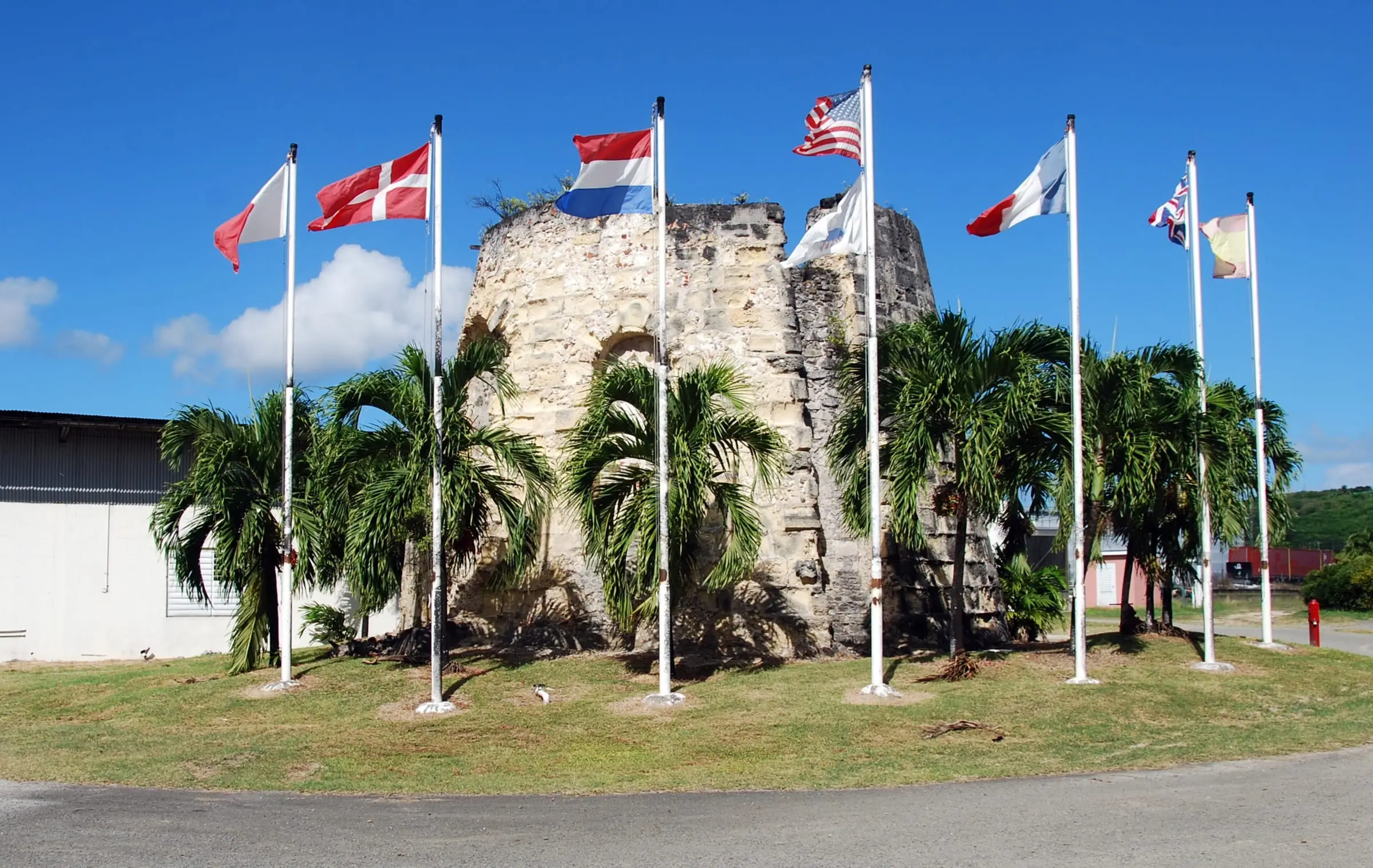 Stone sugar mill ruin at the Cruzan Rum Distillery, ringed by palm trees and flagpoles flying national flags, with a white distillery building at left