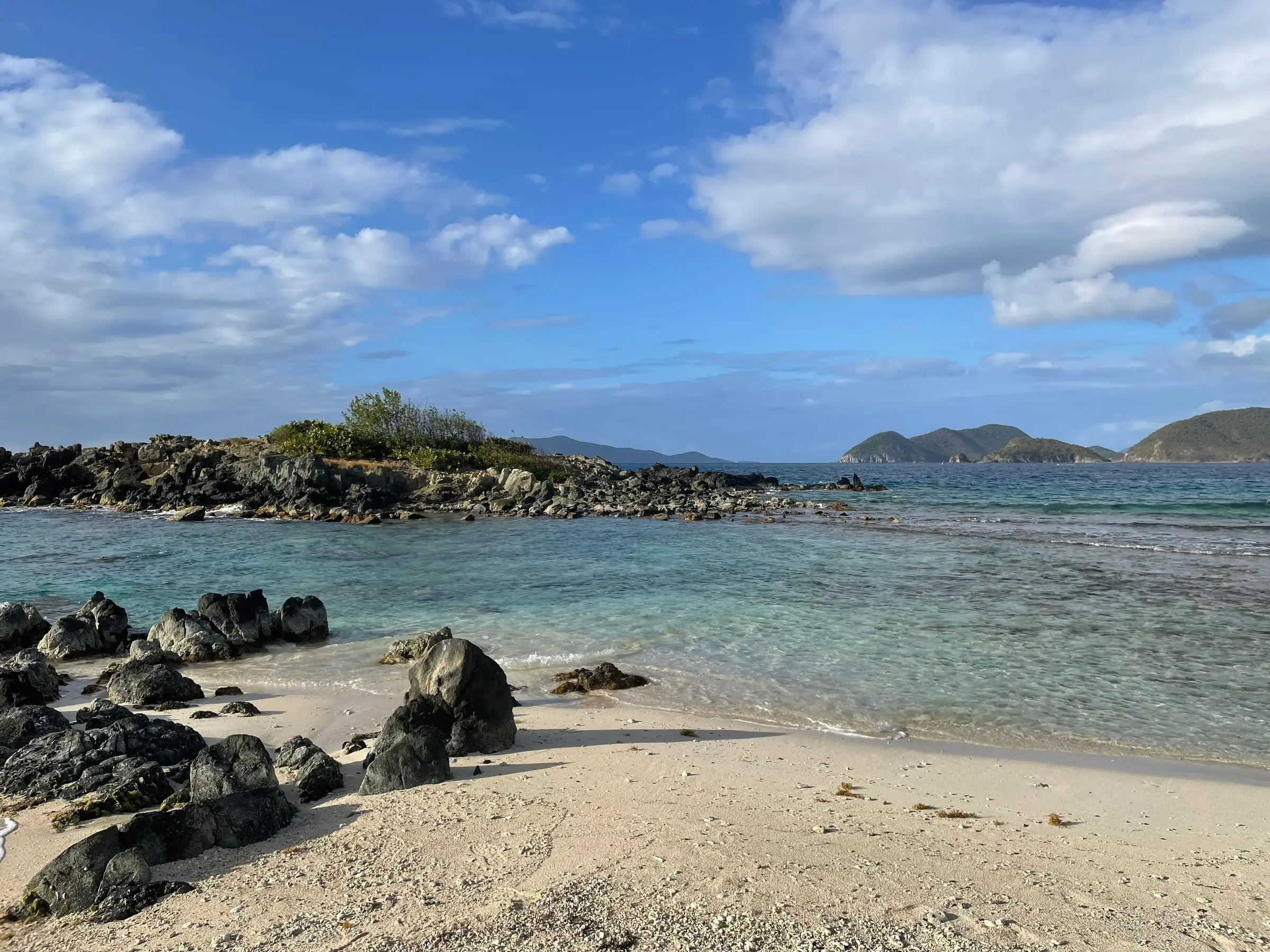 Dark boulders on the white sand of Denis Bay facing the rocky, brush-covered islet of Perkins Cay and green islands on the horizon