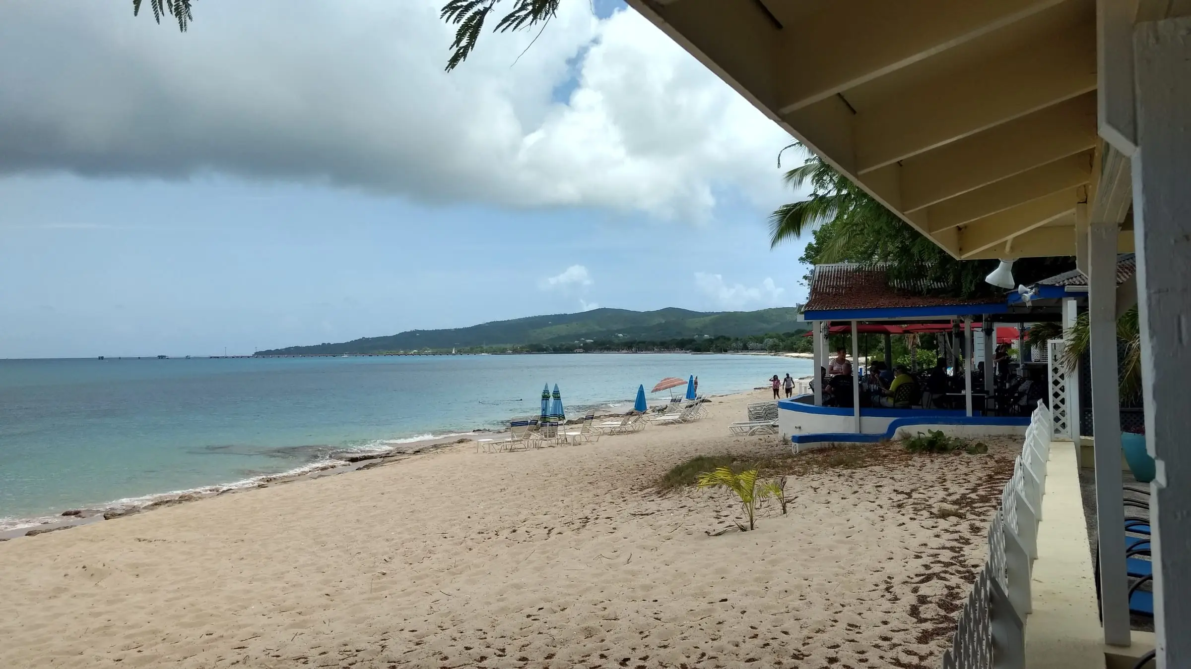 Soft sand of Dorsch Beach curving north toward the Frederiksted Pier and the green hills of the West End, with beach chairs, blue umbrellas and a beachside bar under the palms
