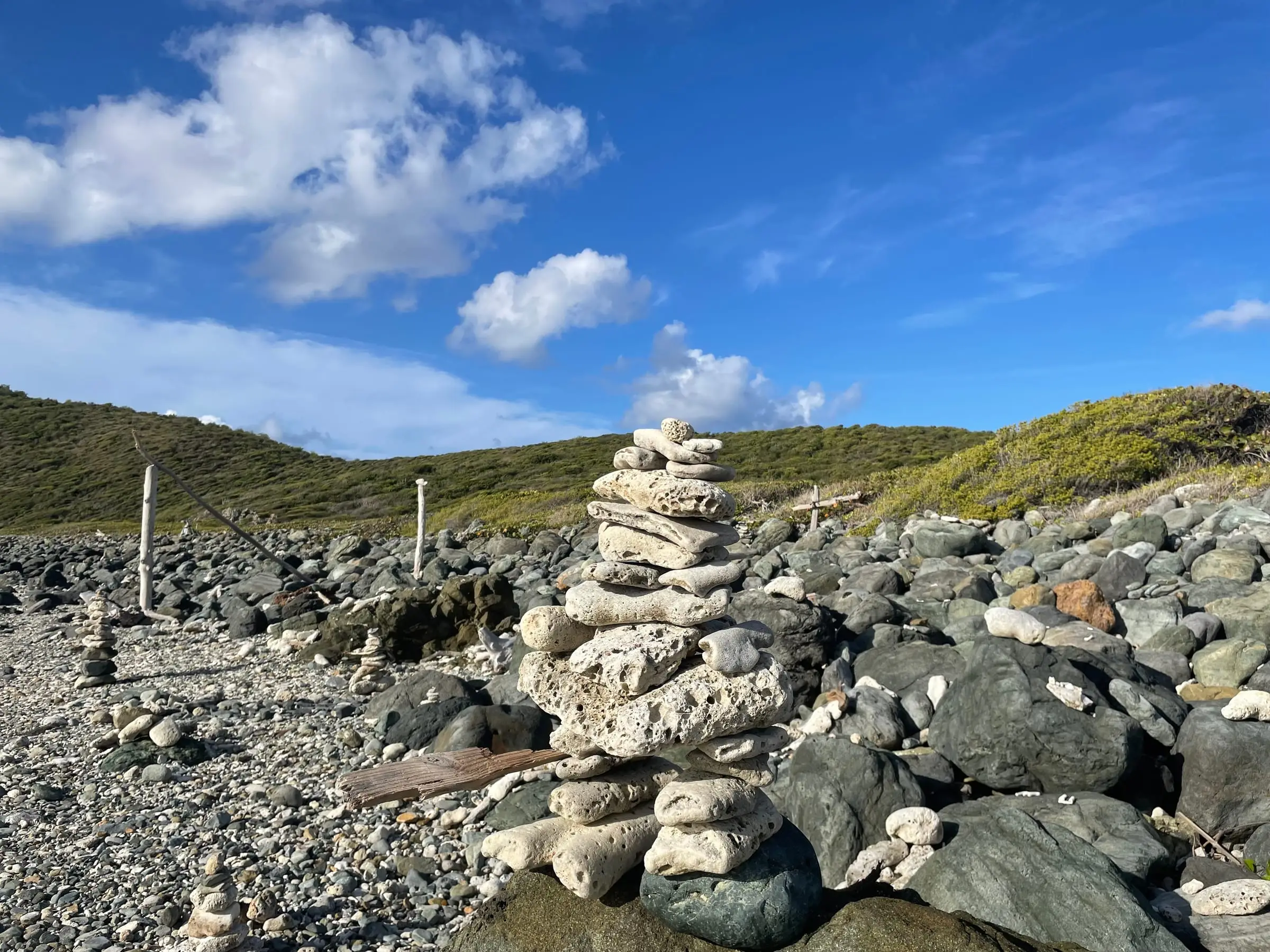 Stacked coral cairn on the rocky shore of Drunk Bay, with driftwood posts, smaller stone stacks and low scrub-covered hills behind