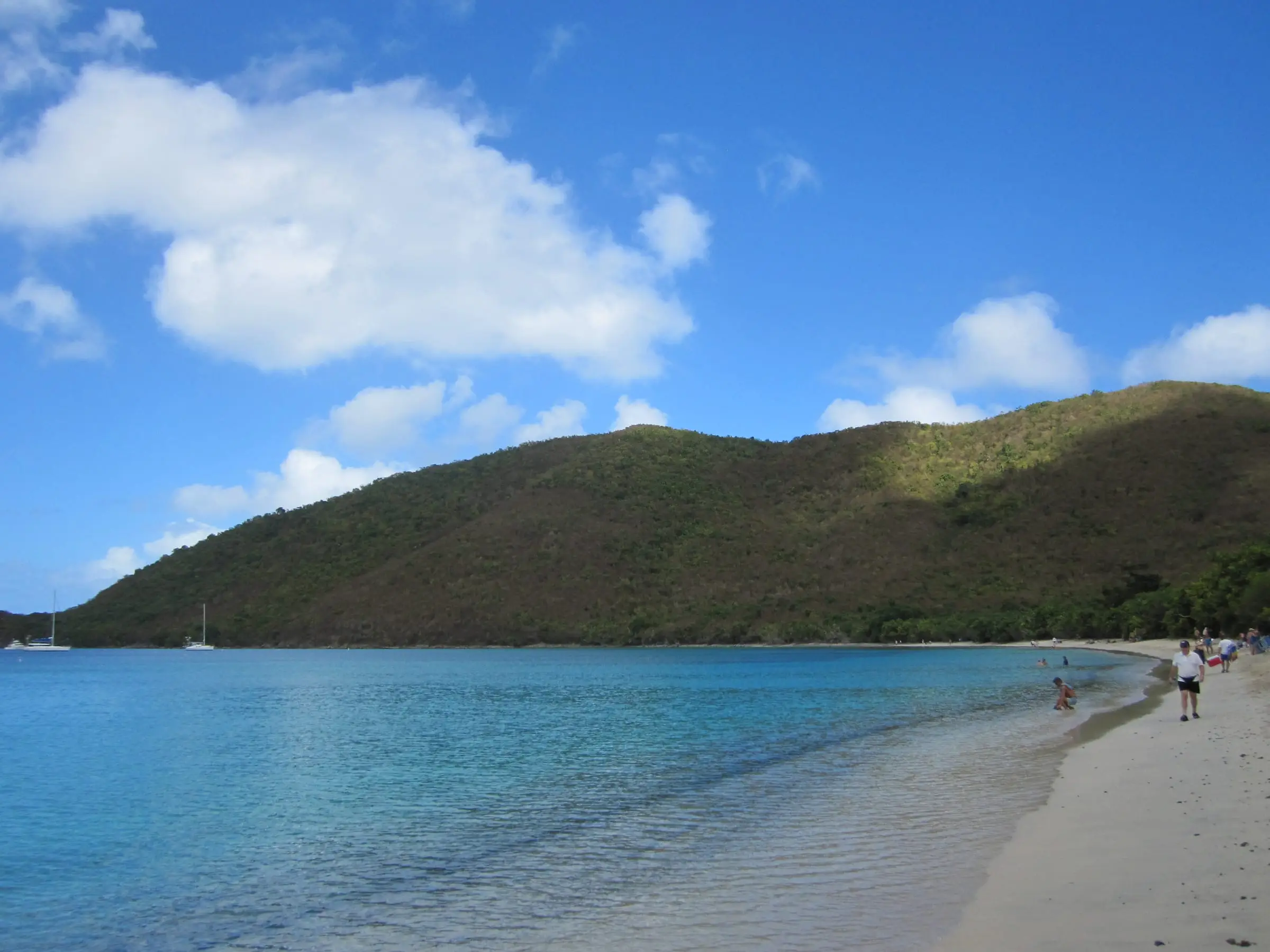 Calm turquoise water along the sandy shore of Francis Bay, with a few beachgoers, sailboats at anchor and a wooded hill rising behind the beach