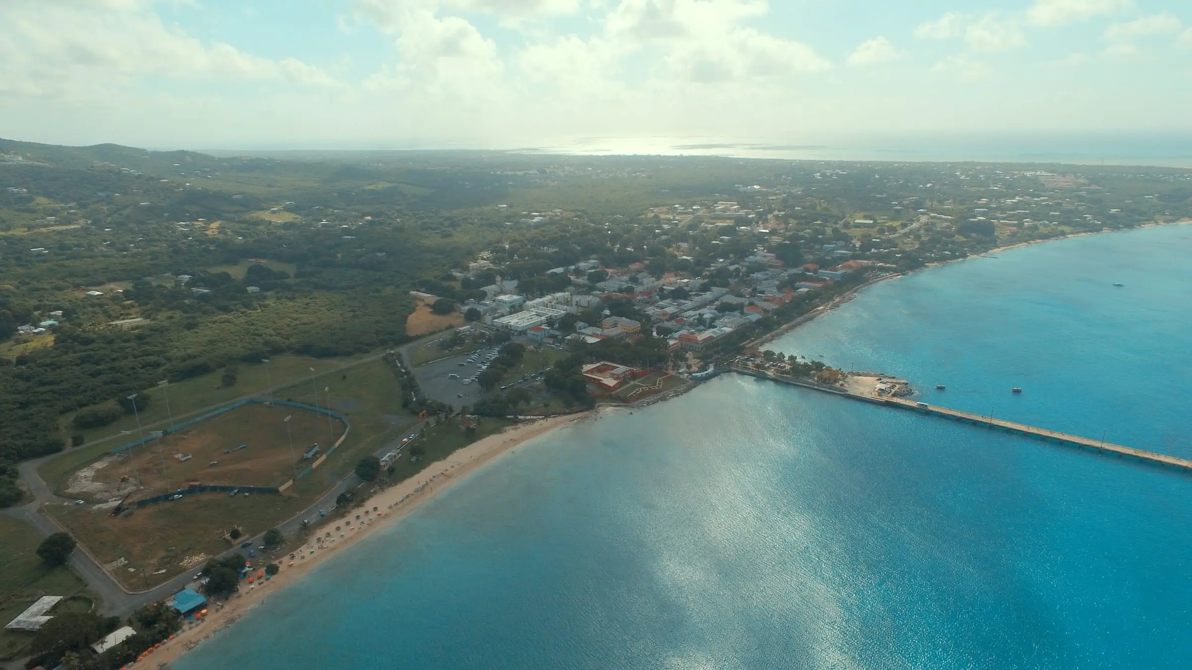 Aerial view of Frederiksted Beach curving north from Fort Frederik and the cruise pier, with umbrellas on the sand, a ballpark behind the beach and the town beyond