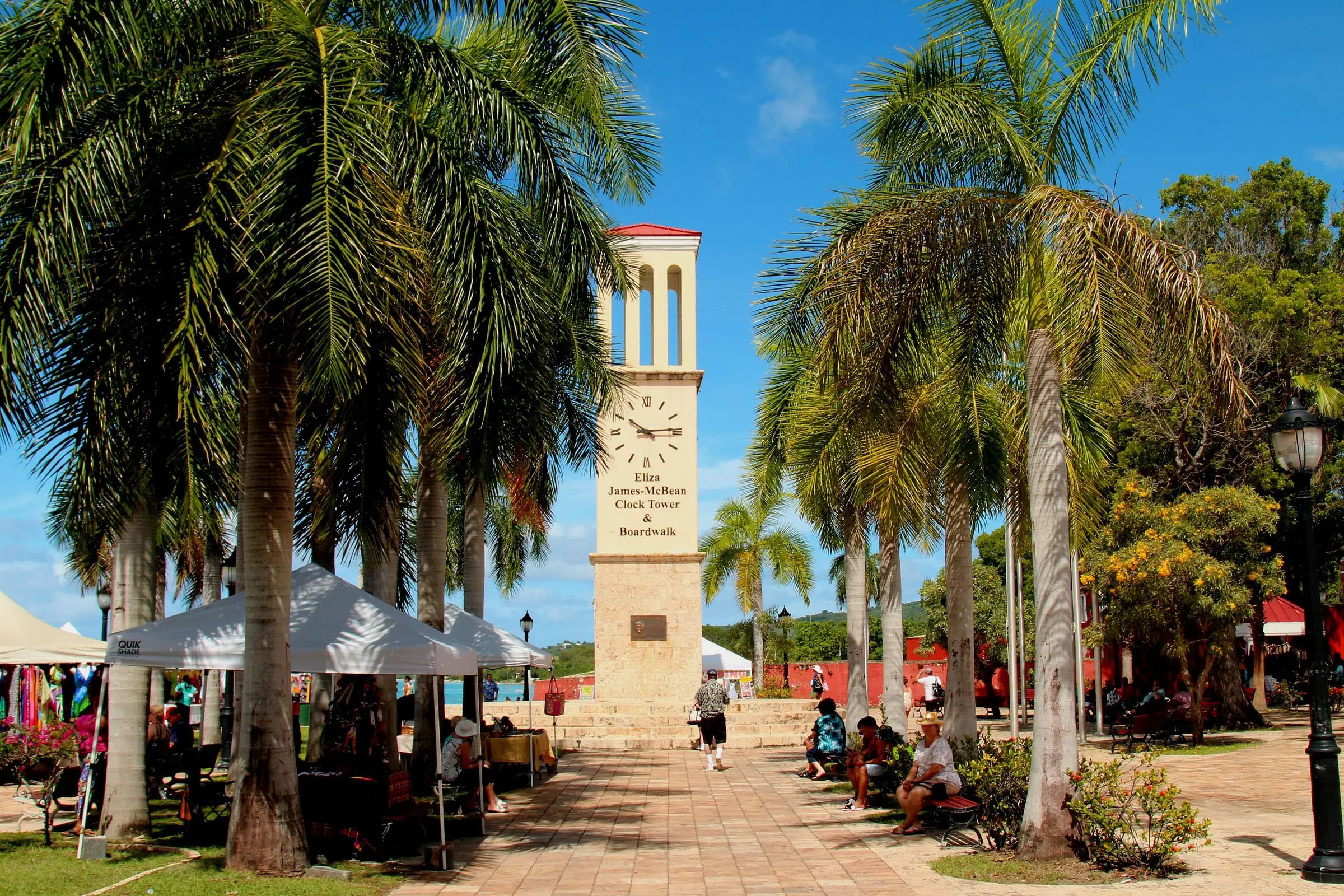 The Eliza James-McBean Clock Tower framed by royal palms on the Frederiksted waterfront, with vendor tents and the red walls of Fort Frederik nearby
