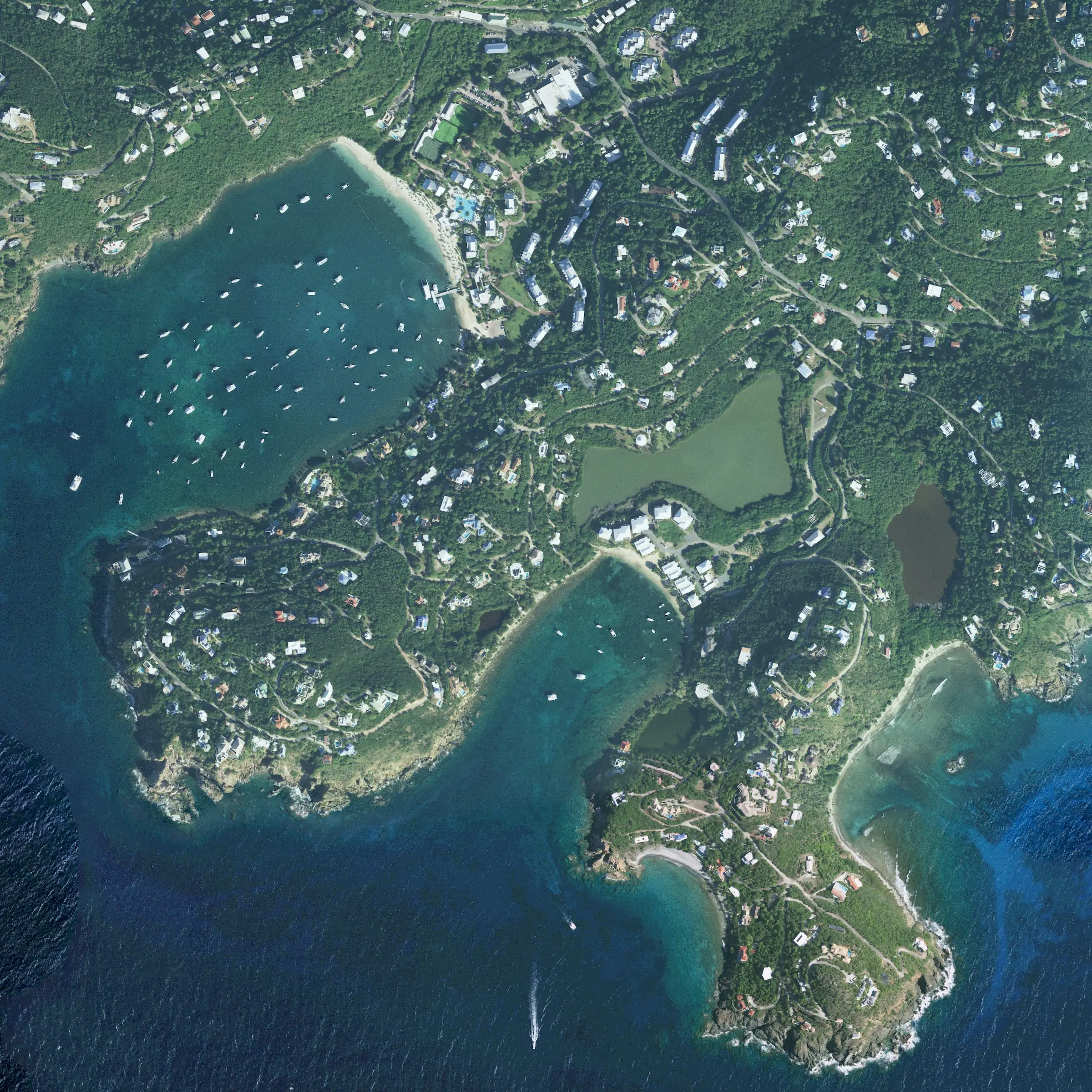 A straight-down aerial view of Great Cruz Bay, with many boats moored off a beach and resort, and the smaller Chocolate Hole bay to the southeast, among hillside homes on St. John