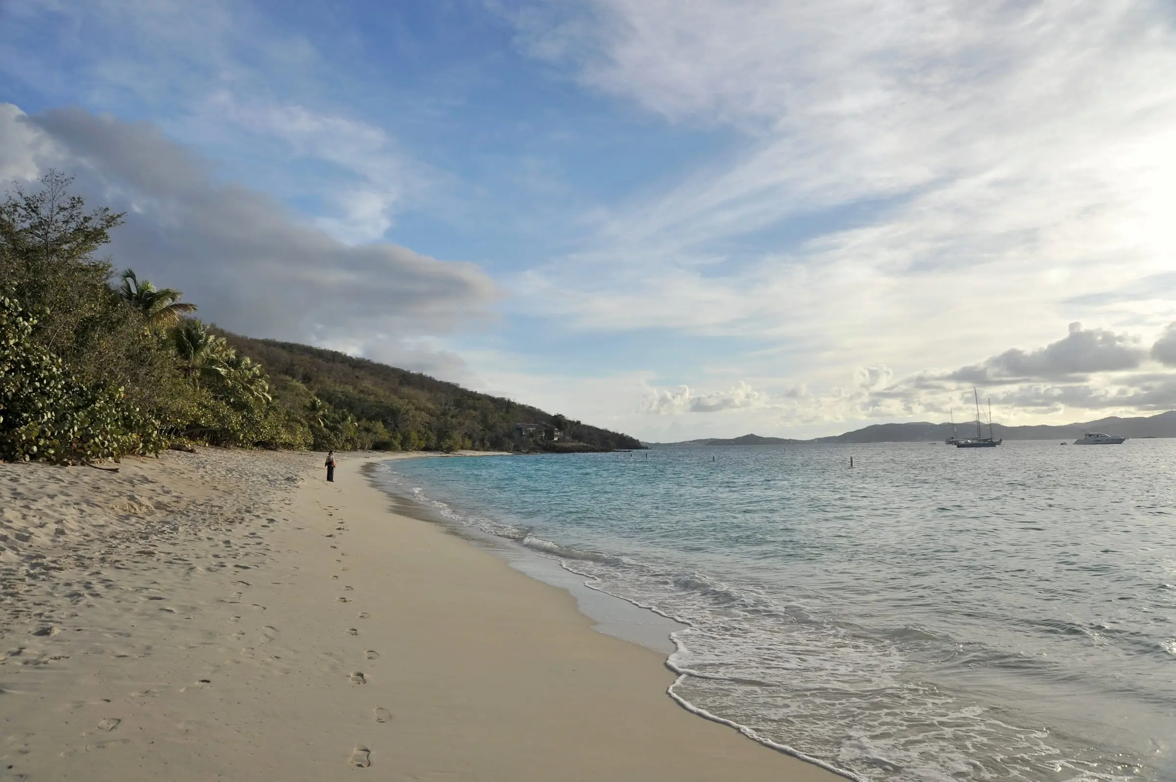 Long sandy beach at Honeymoon Beach with footprints along the waterline, a wooded hillside and sailboats moored offshore