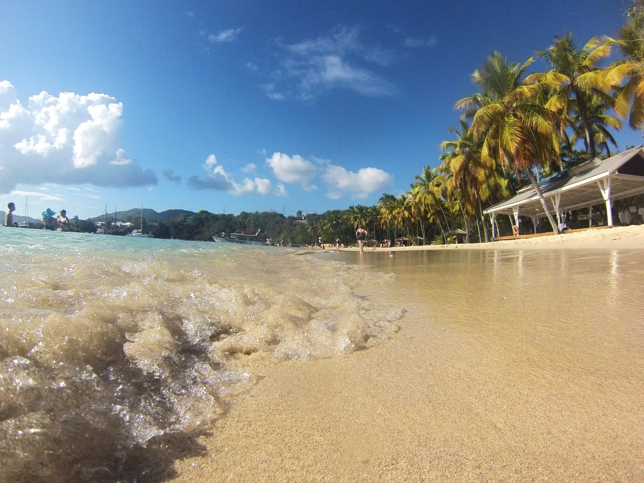 Low view across wet sand and a small breaking wave at Honeymoon Beach on Water Island, with swimmers at left, coconut palms and a beach pavilion along the shore and boats moored in the bay