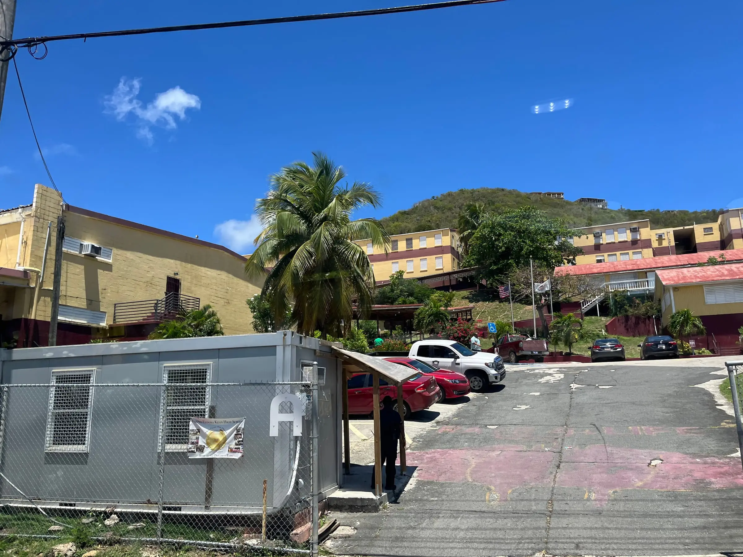 Yellow and maroon buildings of Ivanna Eudora Kean High School climbing a green hillside above palms, flagpoles and the gated entrance drive