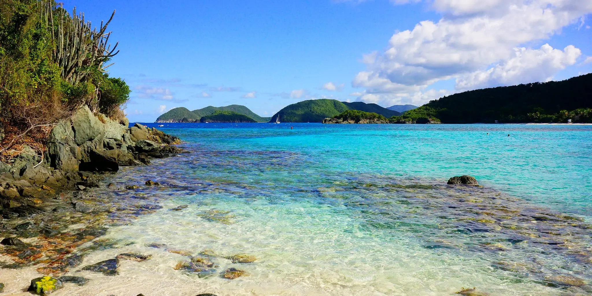 White sand and clear shallow water at Jumbie Beach beside a rocky, cactus-covered point, with green islands and a sailboat offshore and a forested hillside at right