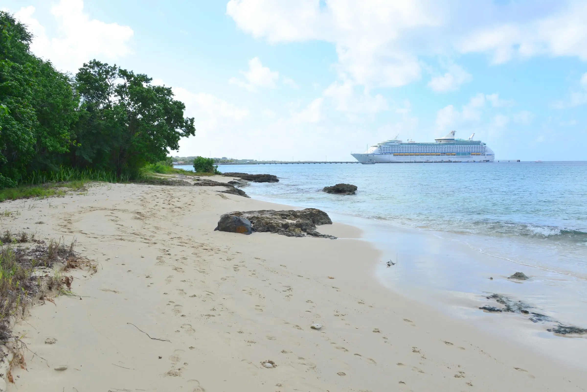 Quiet sand at La Grange Beach with sea grape trees and dark rocks at the water's edge, looking south to a cruise ship docked at the Frederiksted Pier