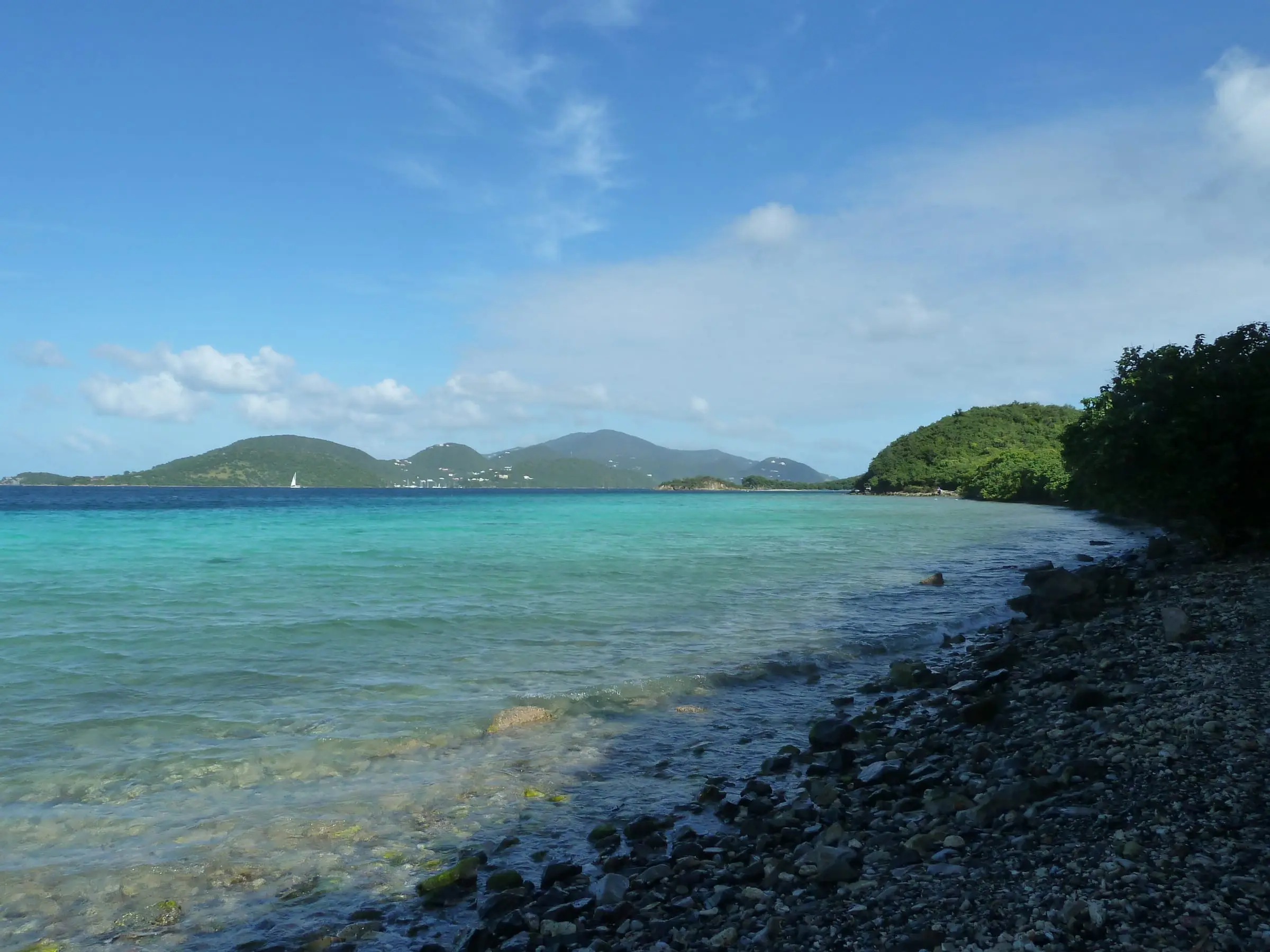 Clear turquoise water along the rocky shore of Leinster Bay seen from the Leinster Bay Trail, with Waterlemon Cay and green hills beyond