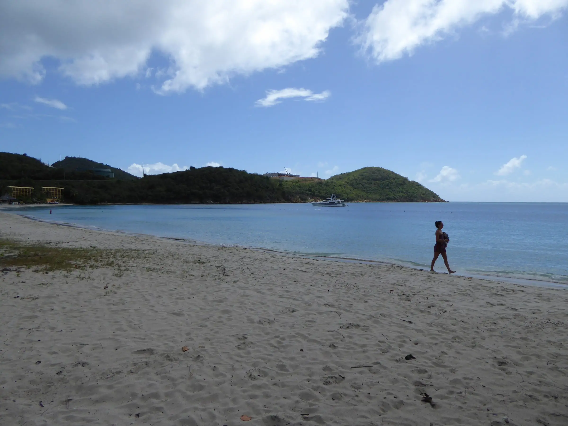 Wide sandy beach curving along the calm water of Lindbergh Bay, with a lone walker at the water's edge, a motor yacht at anchor, a green headland and hotel buildings at the far end of the shore