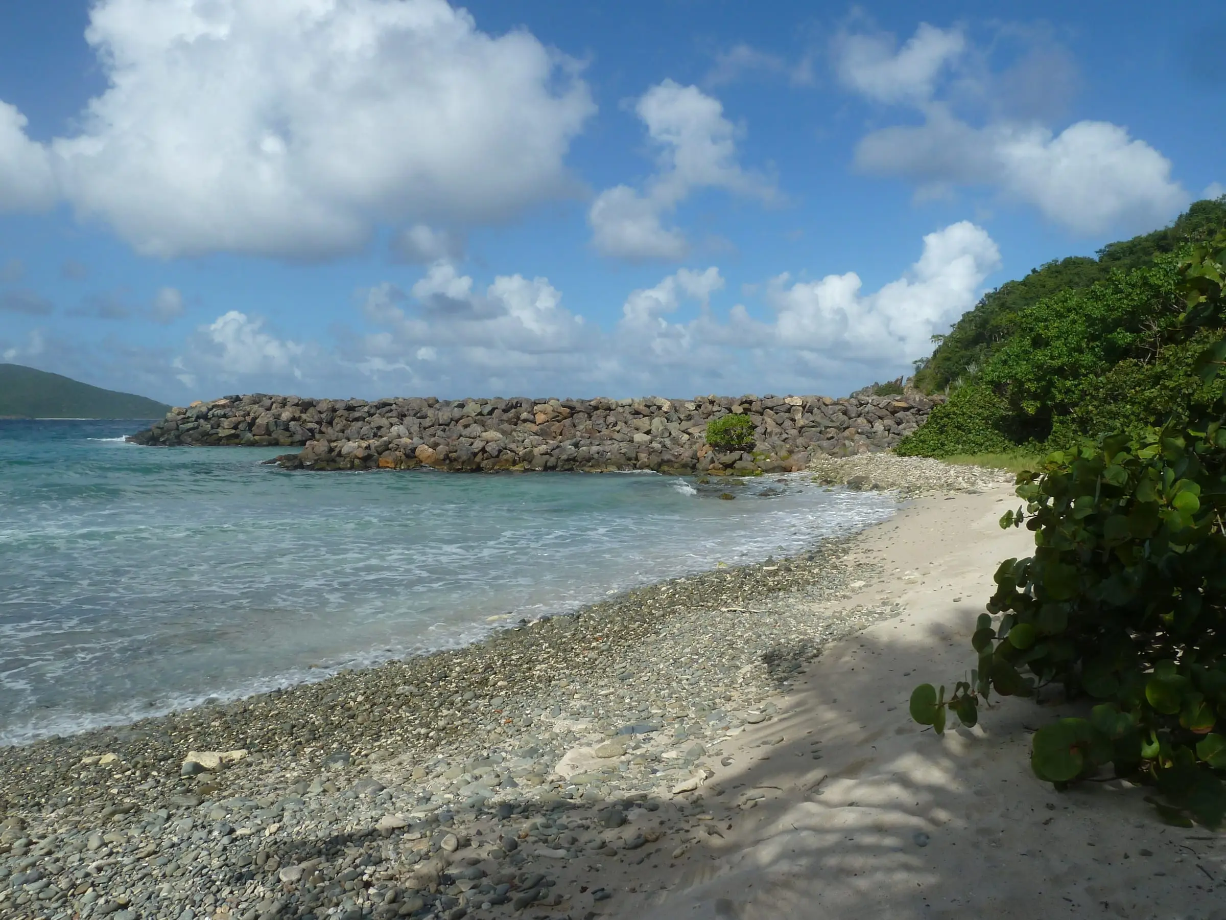 Sand and pebble shore of Mandahl Bay Beach on the north side of St. Thomas, with a rock breakwater and a green hillside