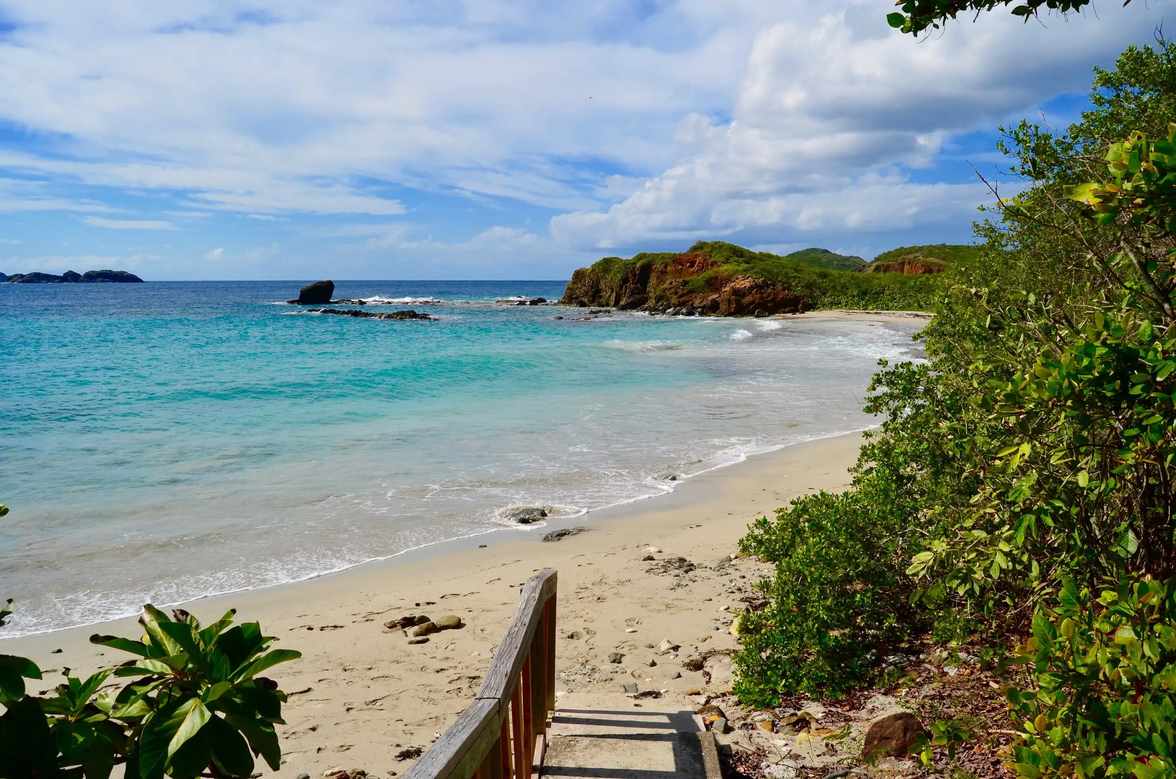 Wooden steps leading down to the sandy curve of Mermaid's Chair beach on the west end of St. Thomas, with turquoise water, offshore rocks and a red cliff headland