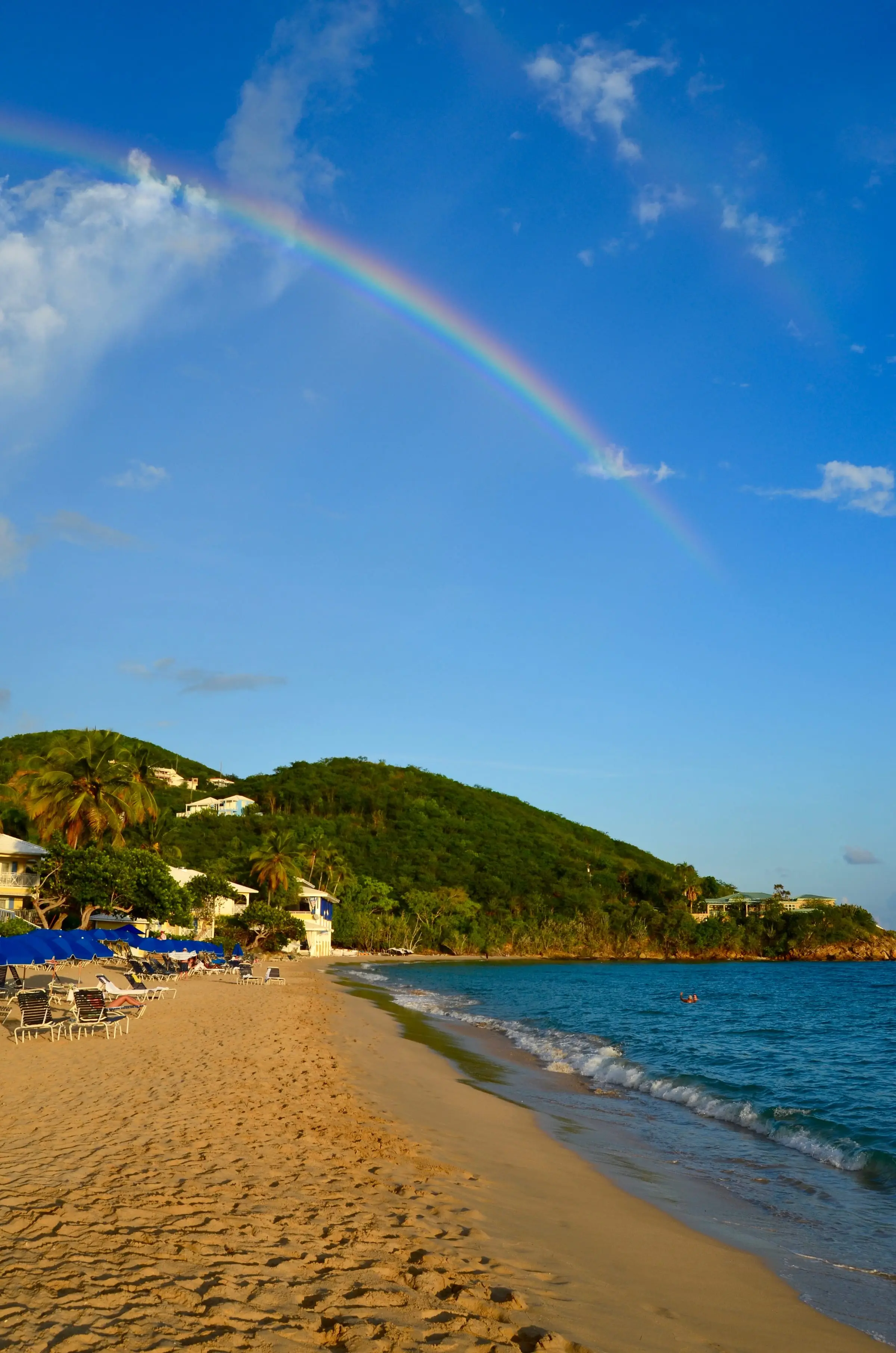Rainbow arching over the golden sand of Morningstar Beach, with beach chairs and blue umbrellas along the shore and a green headland beyond