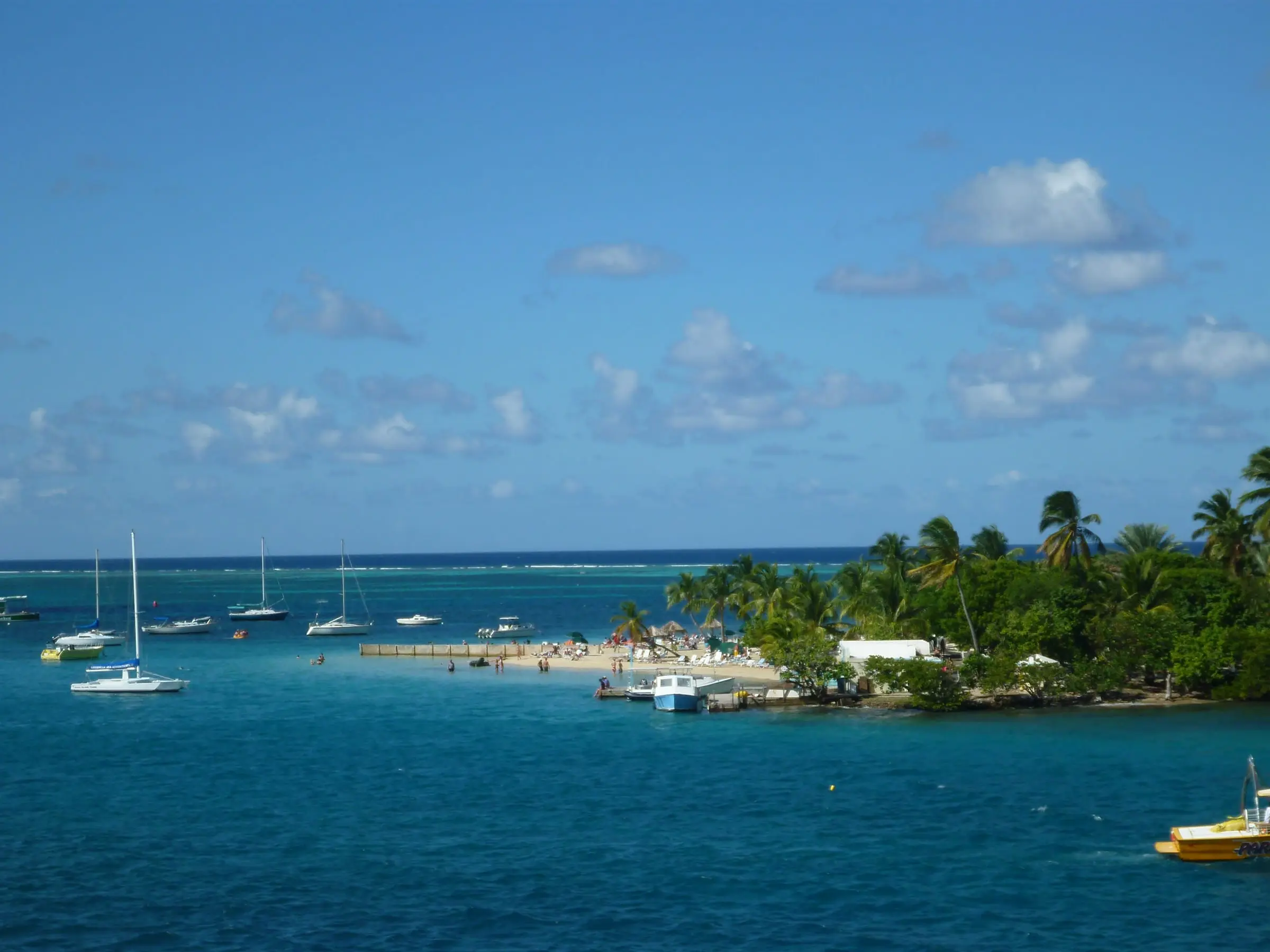 Protestant Cay in Christiansted Harbor, with palm trees, a small beach with umbrellas and swimmers, a dock and sailboats moored in the blue water