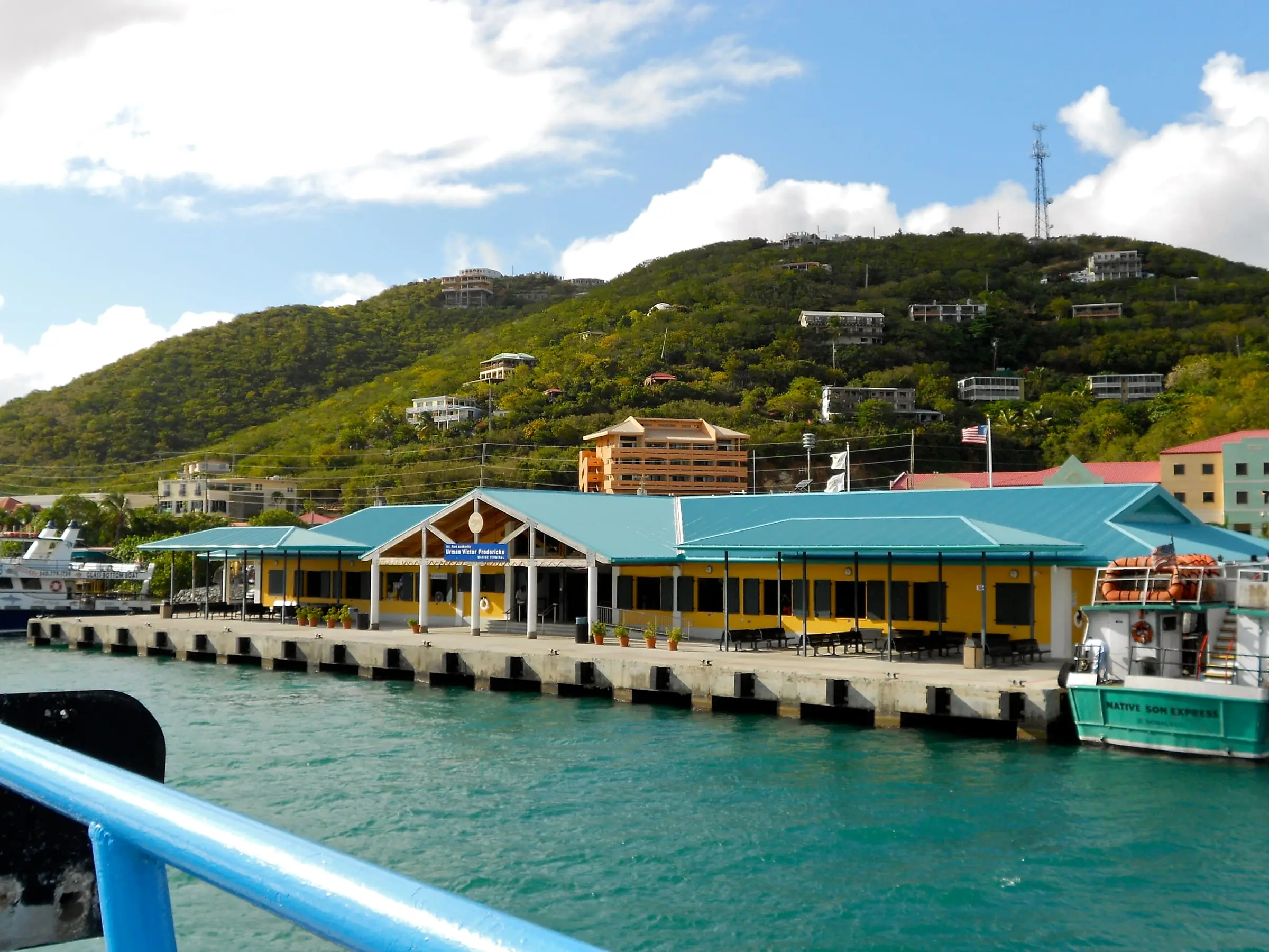 Yellow, teal-roofed Urman Victor Fredericks Marine Terminal on the Red Hook waterfront, seen from an arriving ferry, with homes on the green hillside above