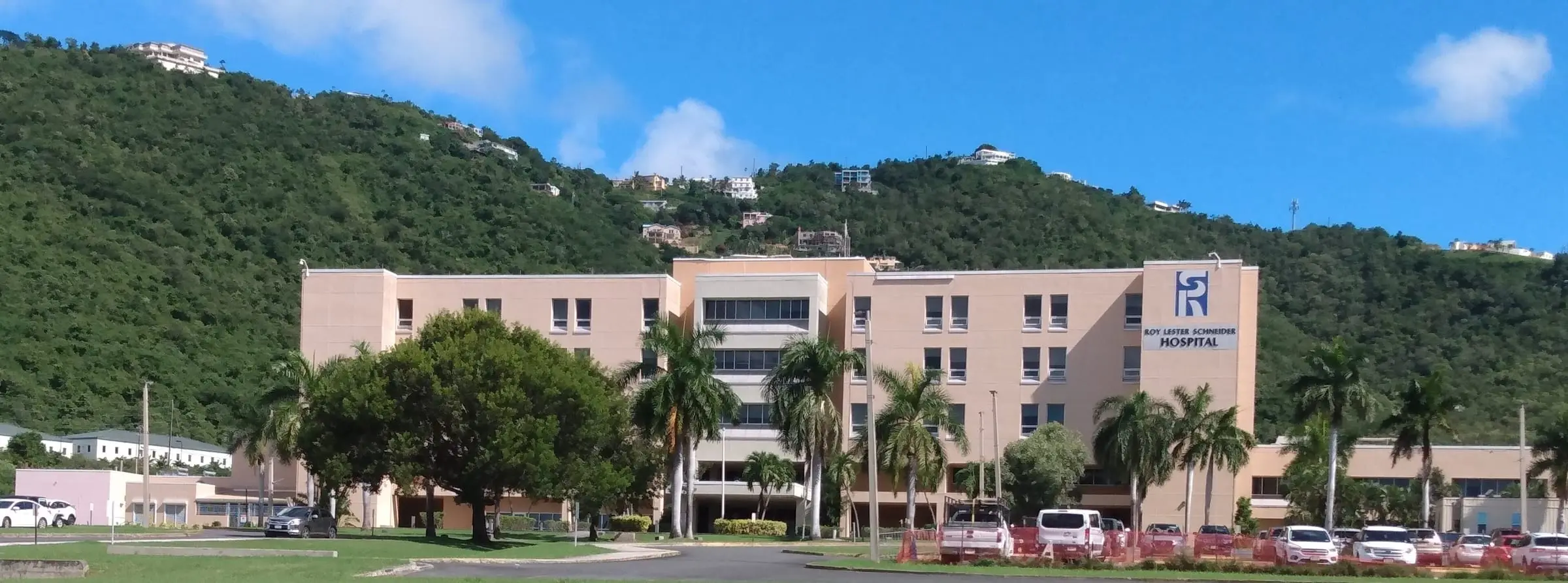 Five-story, peach-colored Roy Lester Schneider Hospital with its name sign, palm trees and a parking lot, below green hills dotted with homes