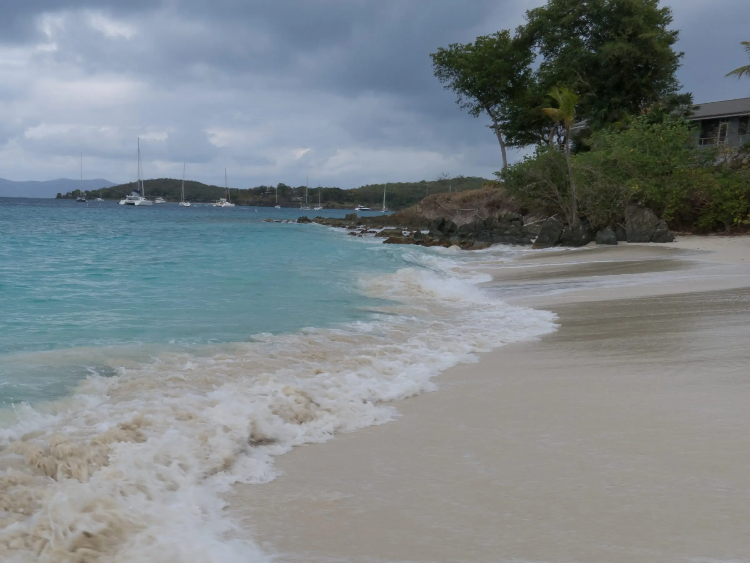 Waves washing onto the white sand of Salomon Beach toward a rocky point, with sailboats moored offshore under a cloudy sky