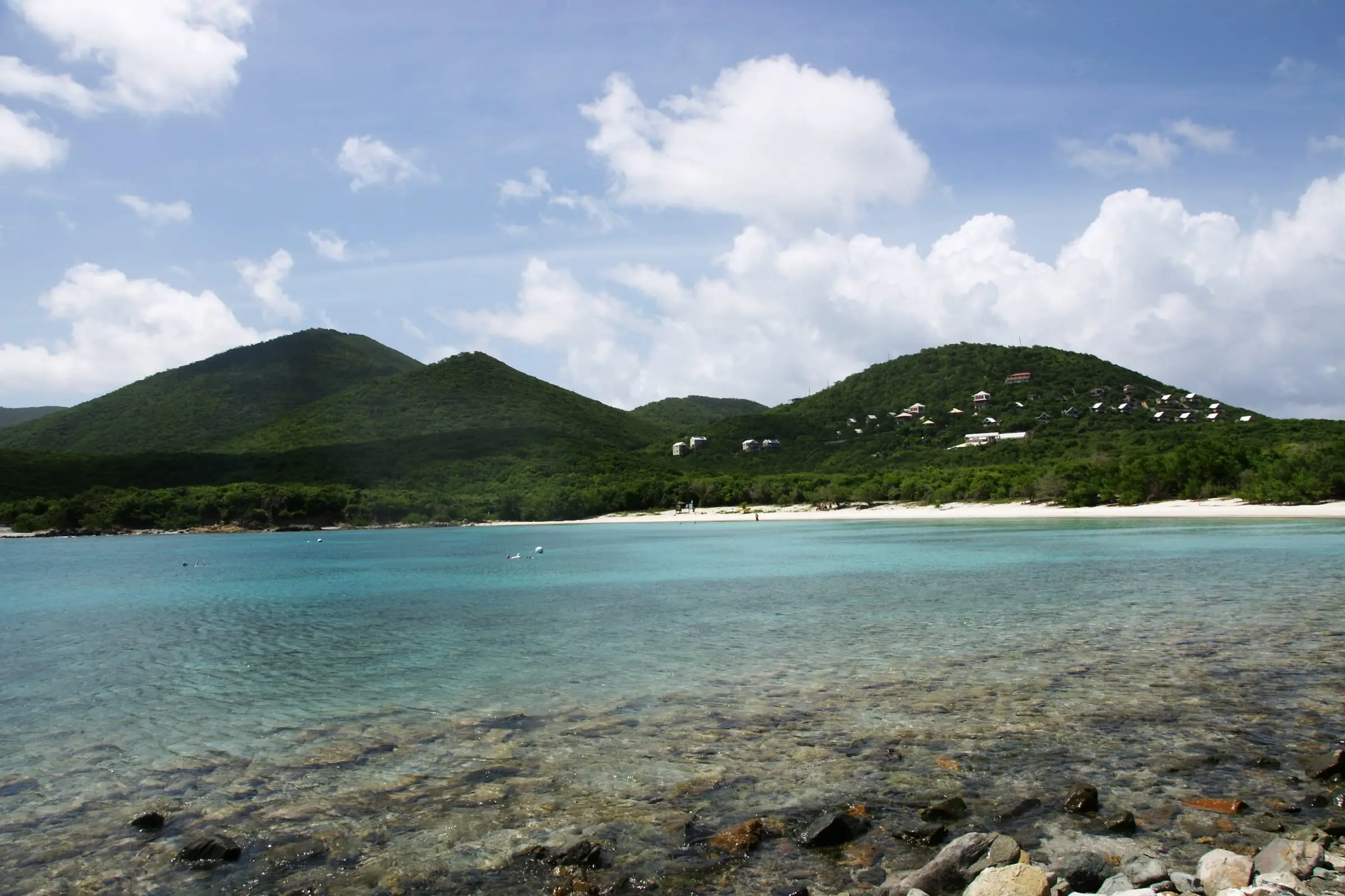 Salt Pond Bay seen from the rocky shore across the bay, with clear turquoise water, snorkelers near the buoys, the white beach beyond and small cabins on the wooded hillside above