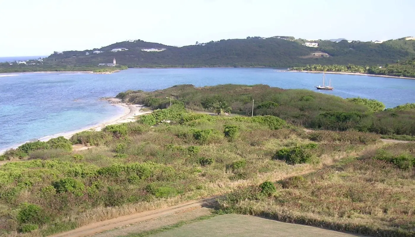 Scrub-covered point and sandy beach at the Columbus landing site on Salt River Bay, seen from the park's visitor center, with a sailboat moored in the bay and hills beyond
