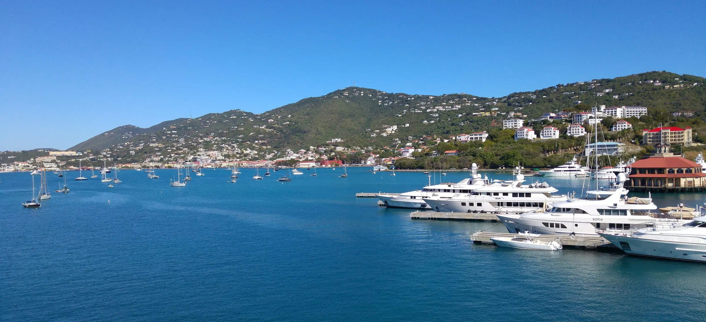 Megayachts at the docks of Yacht Haven Grande beside its round red-roofed pavilion, with sailboats in St. Thomas Harbor and Charlotte Amalie on the hills beyond